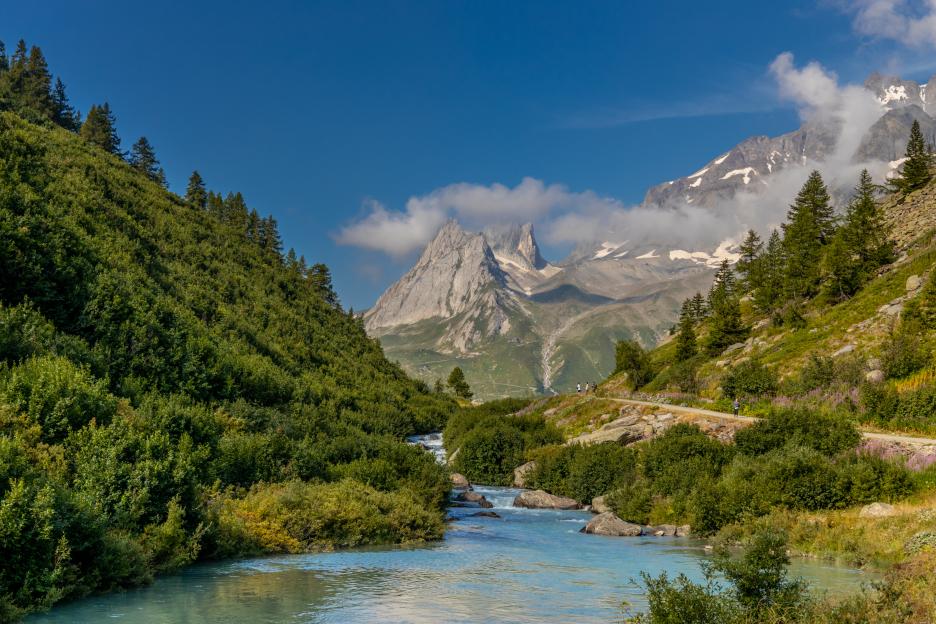 Val Veny scenic landscape in Courmayeur valley, Italy, with alpine peaks, a blue river, and hikers on the Tour du Mont Blanc route.