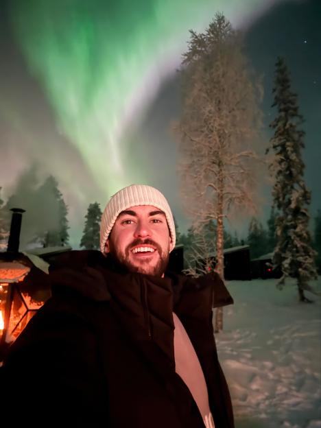 A man smiling with snow on his beard and eyebrows, wearing a white hat and a black jacket, against a backdrop of green aurora borealis, snow-covered trees, and distant cabins.