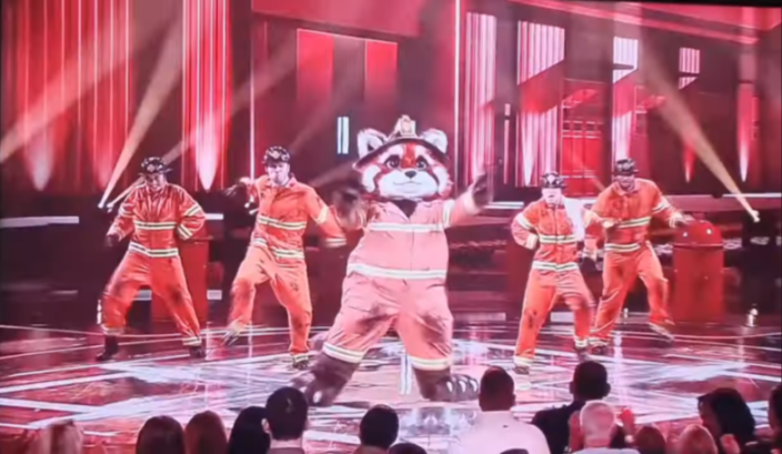 A red panda mascot dressed as a firefighter performing on stage with four other dancers in firefighter uniforms.