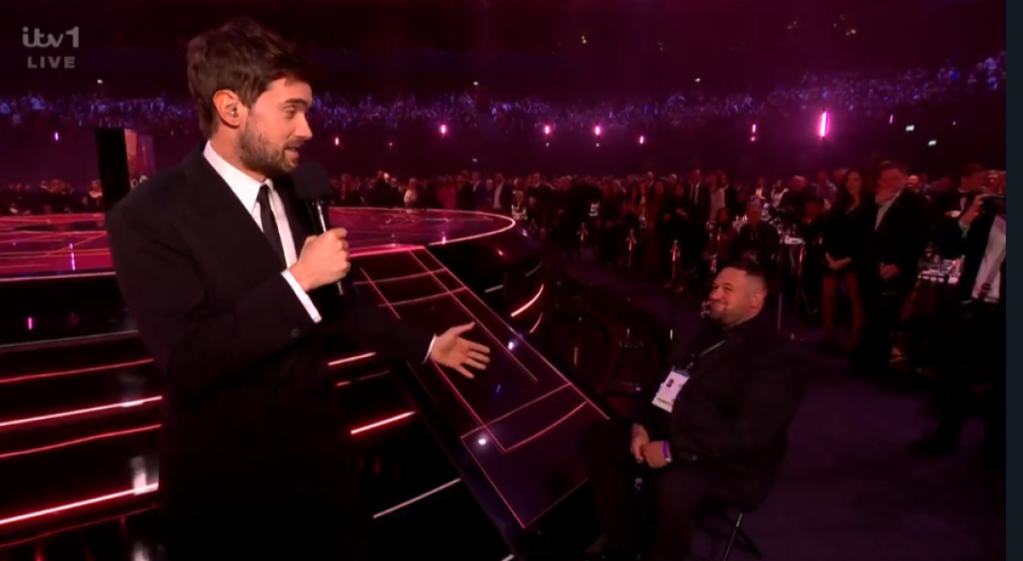 A man in a suit speaking into a microphone on stage, gesturing towards another man seated on a chair.