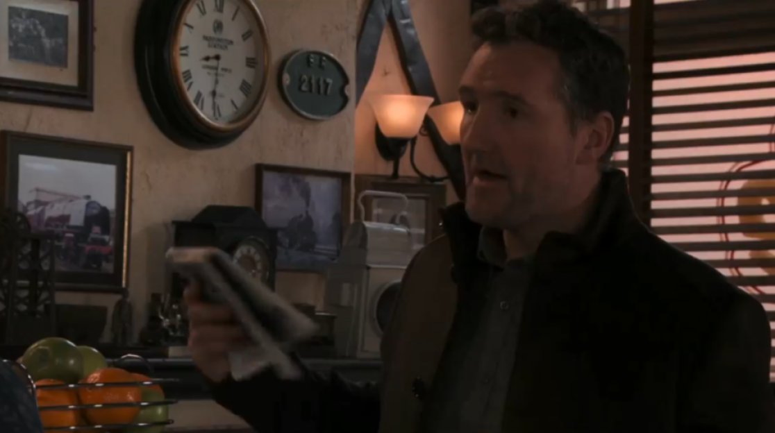 A man in a dark jacket talking, holding an object, in a room decorated with clocks and framed train pictures.