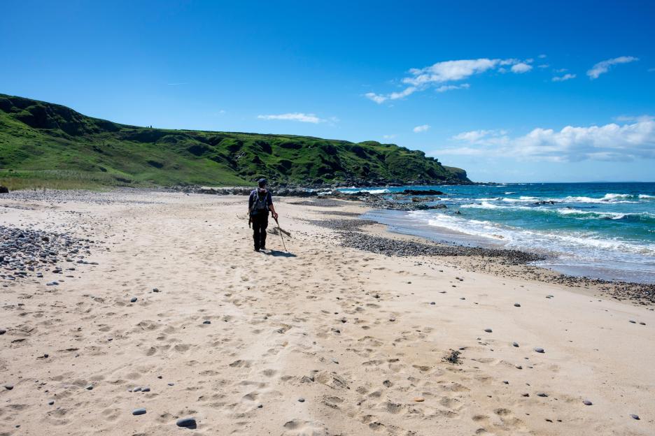 A person with a backpack and a walking stick walks on a sandy beach towards the ocean, with green hills in the background.