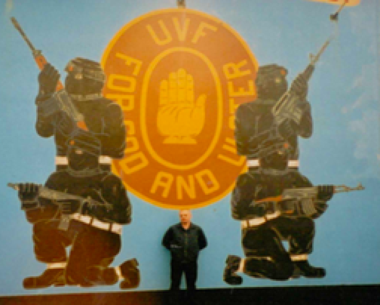 Nigel Bromage standing in front of a mural depicting four armed figures and the UVF logo.