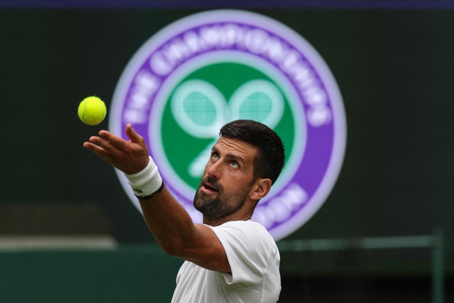 Novak Djokovic prepares to serve, tossing the tennis ball into the air, with the Wimbledon logo in the background.