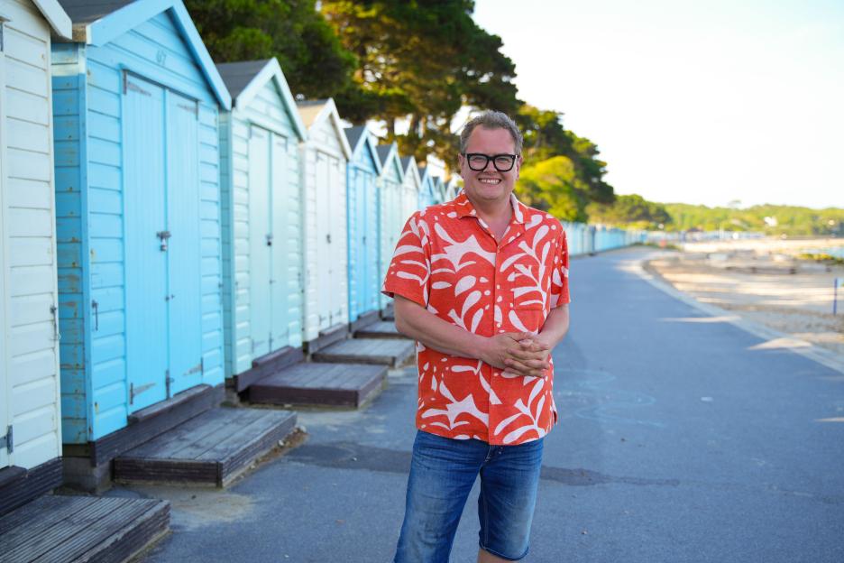 Alan Carr stands in front of a row of blue and white beach huts.