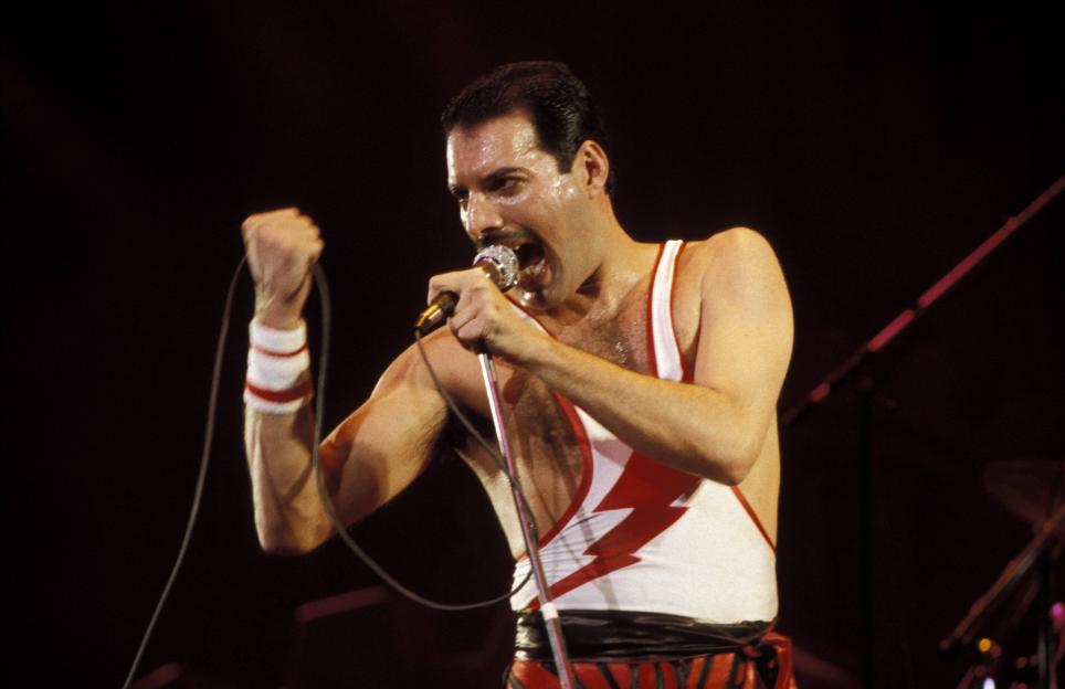 Freddie Mercury performing live on stage, wearing a white singlet with a red lightning bolt design, holding a microphone.