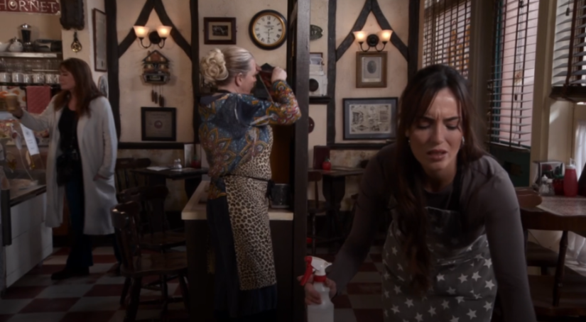 Three women in a café, one holding a drink, another tidying up, and a third looking distressed while holding a spray bottle.