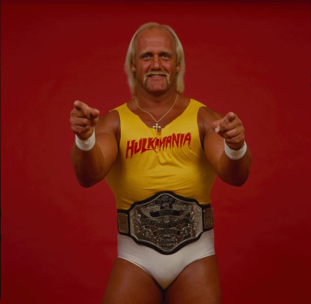 Hulk Hogan in a yellow "HULKAMANIA" tank top and championship belt points at the camera.