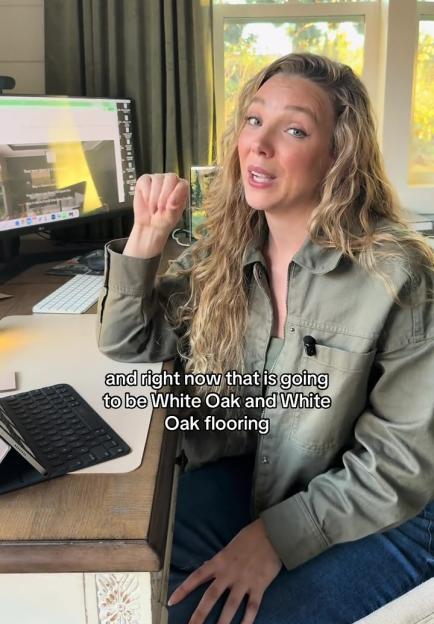 A woman sitting at a desk with a computer monitor showing a home design program, gesturing while speaking.
