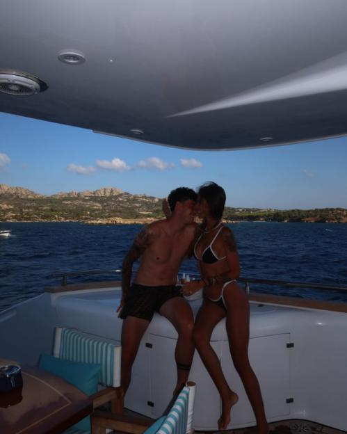 A man and a woman in swimwear kiss on a yacht with mountains in the background.