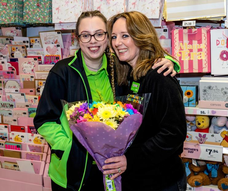 Sian, with glasses and braided hair, and her mother Kuia, both employees, pose with a bouquet of flowers in a supermarket aisle filled with Mother's Day gifts.