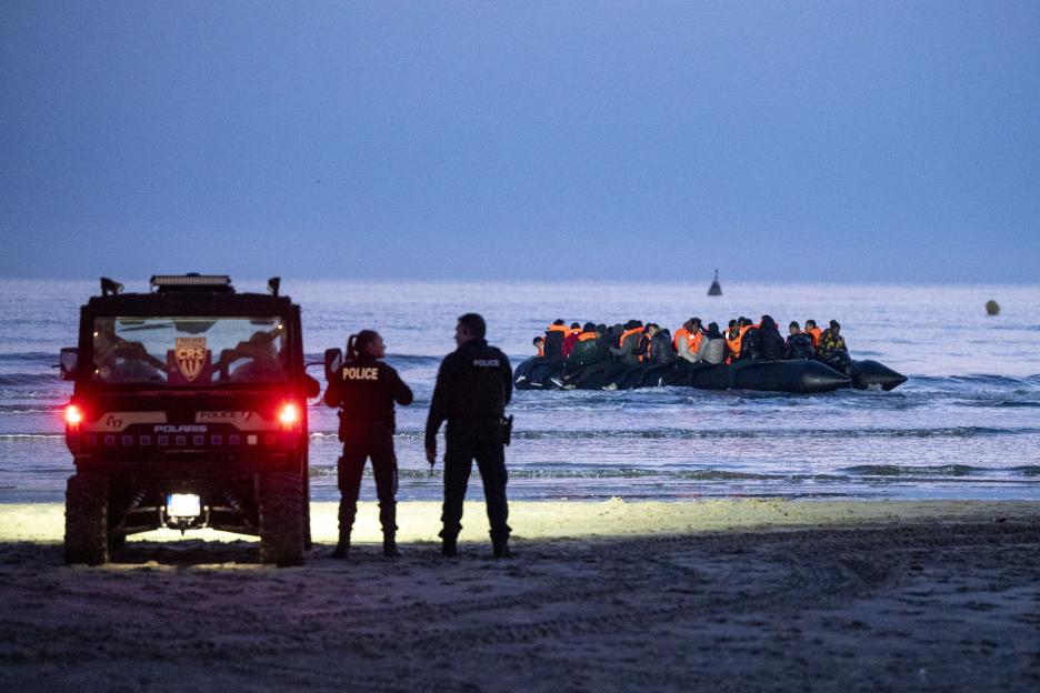 Two police officers stand on a beach next to a police vehicle as migrants attempt to depart on an inflatable boat.