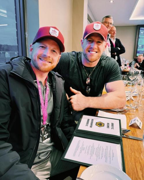 Two men in matching burgundy baseball caps smiling, with a menu for a football match in front of them.