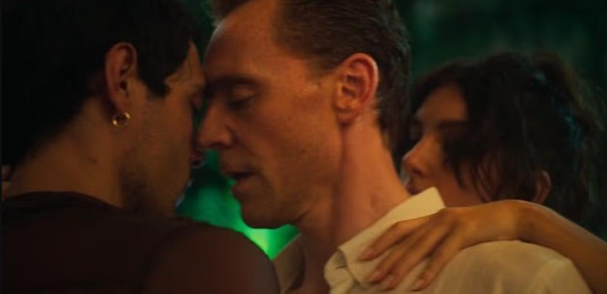 Three people close together, two men with foreheads touching and a woman embracing one of them.