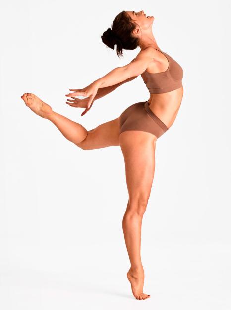 A woman with dark hair in a bun, wearing a nude bra and underwear, poses like a dancer with one leg extended behind her and her arms outstretched.