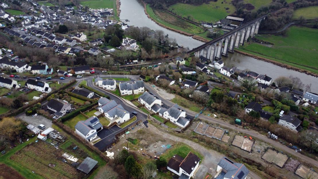 Aerial view of an abandoned new-build estate with some homes completed and others still foundations, alongside a river and a railway bridge.