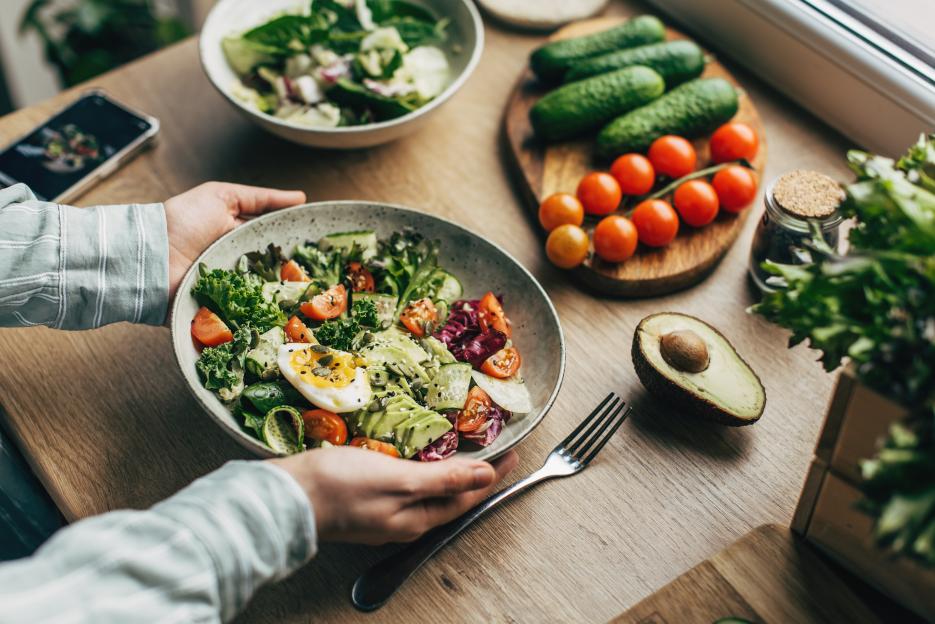 A person holding a bowl of salad with avocado, tomatoes, cucumber, seeds, and a boiled egg.