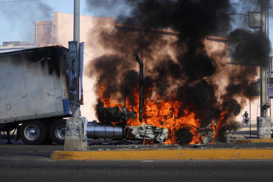 A truck burns with large flames and black smoke on a street.