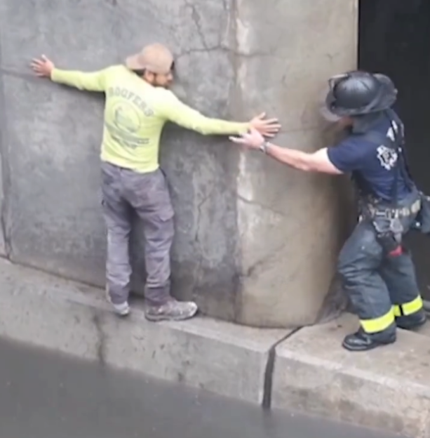 A man in a yellow shirt and a firefighter in a blue uniform are helping each other stand on a flooded street.