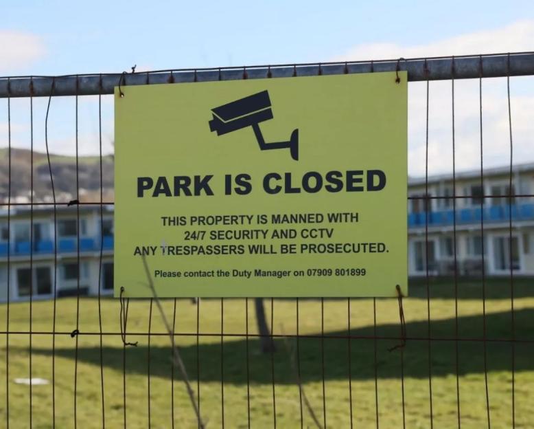 A "PARK IS CLOSED" sign on a fence with a building and grass in the background.