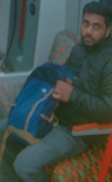A man with a beard, wearing a dark jacket, sitting on a train seat while holding a blue backpack.
