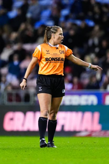 Tara, a St Helens player, officiating a rugby match between Wigan Warriors and St Helens in an orange referee shirt, black shorts, and black socks, with a crowd and "GRAND FINAL" sign in the blurred background.