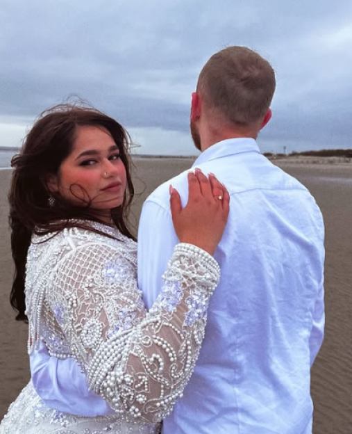 A woman in an ornate white dress with pearls and lace looks at the camera while holding the arm of a man looking away from the camera.