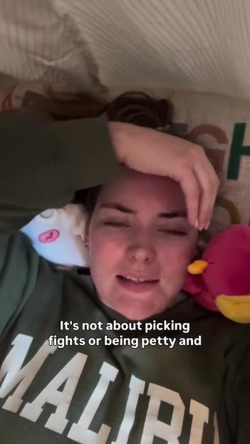 A woman in a green sweatshirt with "MALIBU" written on it looks at the camera with her hand on her forehead and a pink plush toy beside her.