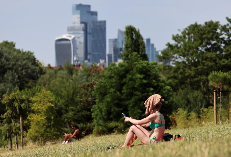 A person in a green bikini and a T-shirt on their head uses a phone in a park with distant city skyscrapers.