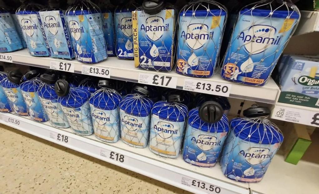 Aptamil baby formula on a store shelf, with security netting and prices displayed.