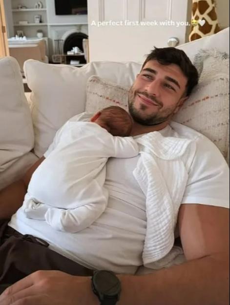 Tommy Fury smiling while holding his newborn baby against his chest.