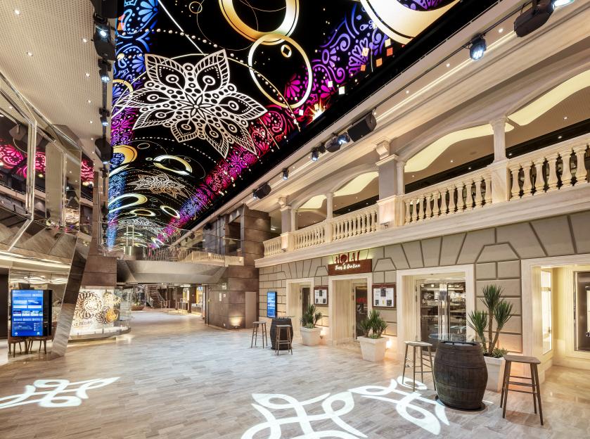 A long, colorful digital ceiling displaying mandala-like patterns and circles in pink, purple, blue, and gold, above a cruise ship's grand promenade featuring a restaurant, shops, and lounge areas.