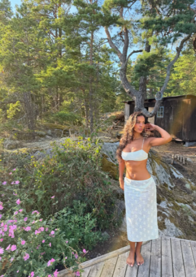 A woman in a white bikini top and white floral sarong standing on a wooden deck in a forest.