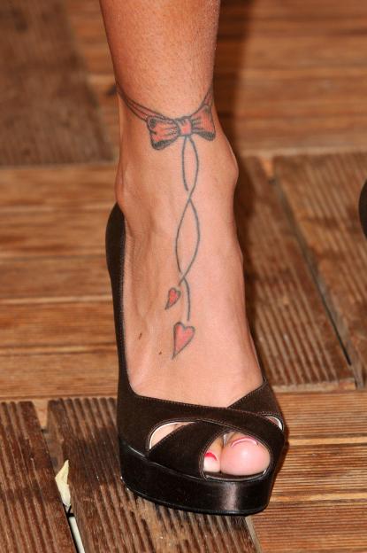 A woman's ankle with a tattoo of a red bow and two hearts, wearing a black peep-toe platform heel.