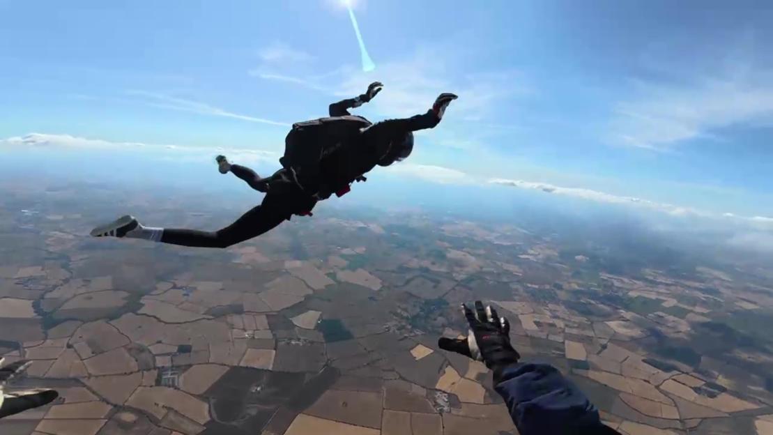 A skydiver freefalling with another skydiver's gloved hand visible in the foreground.