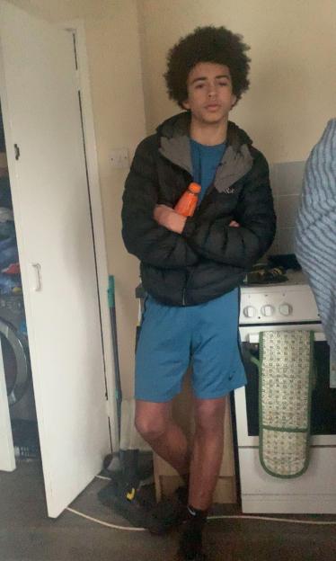Ta-Shay Canoville, a young man with dark, curly hair, in a black jacket, blue shirt and shorts, standing with a bottle in his hand.