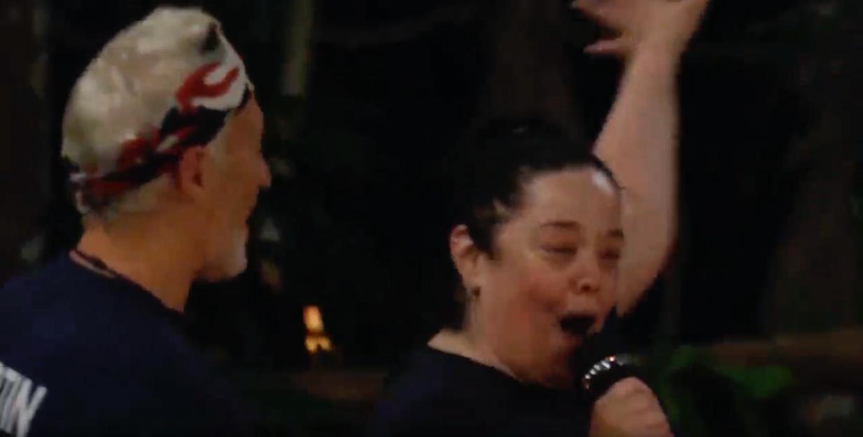 Screenshot from "I'm a Celebrity...Get Me Out of Here!" showing a woman with dark hair singing into a microphone next to a man with white hair and a white and red patterned cap.