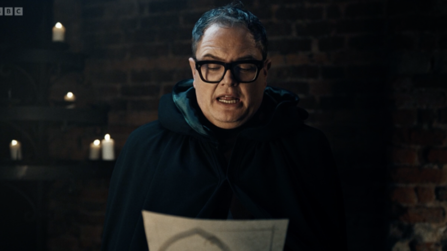 Alan Carr in a dark cloak and glasses reading from a paper, with lit candles behind him.
