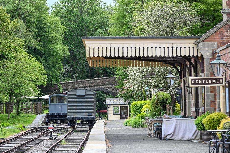 A historic train station with a train car on the tracks, surrounded by lush green trees, with a sign reading "GENTLEMEN."
