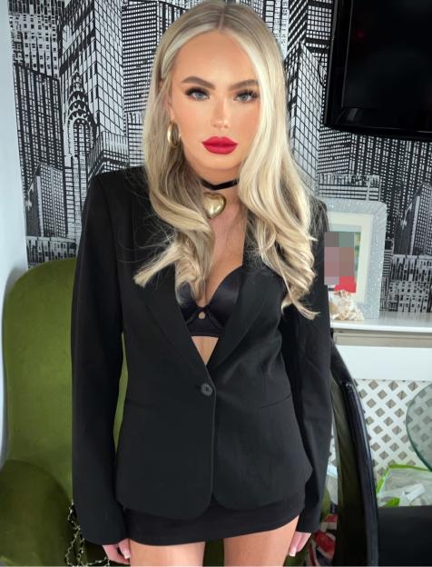 Mia O'Brien, a woman with long blonde hair and bright red lipstick, looks directly at the camera wearing a black blazer over a black bra and a black skirt, in front of a cityscape wallpaper.