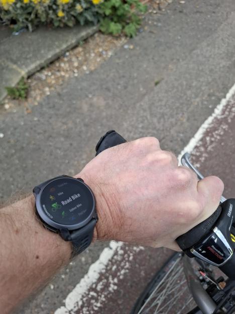 A person's arm on a bicycle handlebar with a smartwatch displaying activity options, including "Road Bike" highlighted.