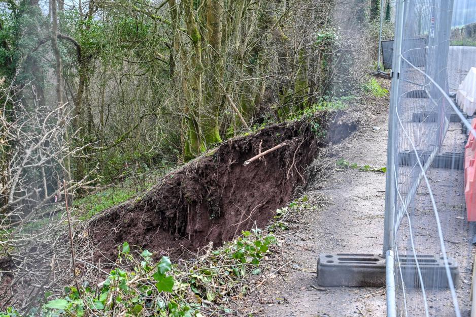 A large landslide has closed a road next to a forest, leaving a vertical drop beside the asphalt, with construction fencing blocking off the dangerous area.