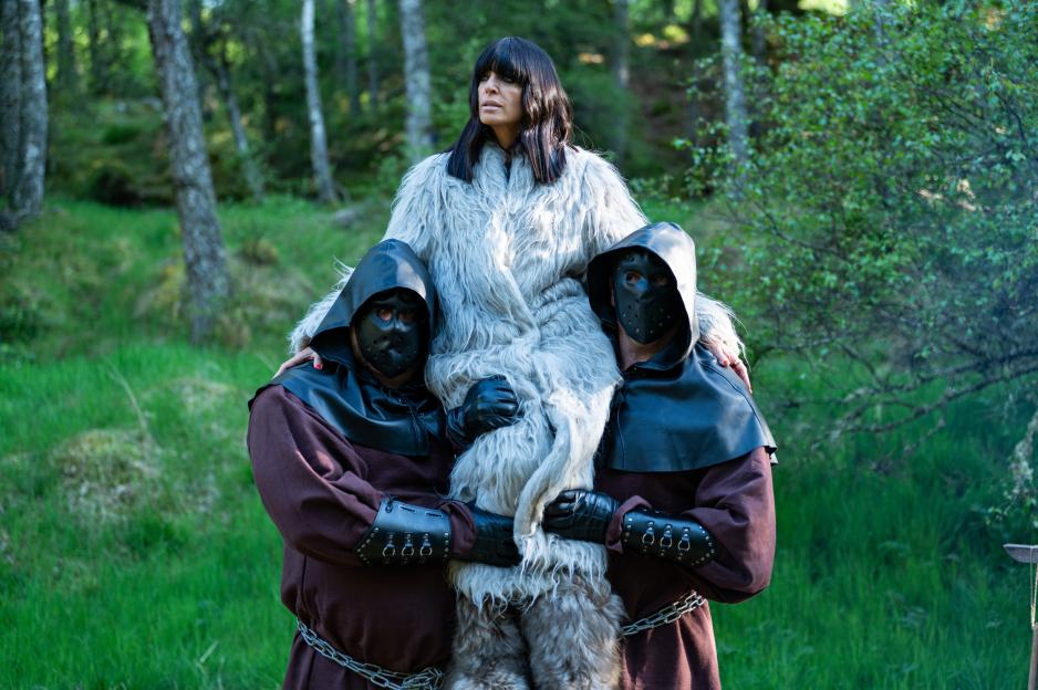 Claudia Winkleman, in a fur coat, is carried by two masked figures.