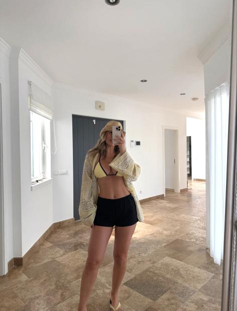 A woman takes a mirror selfie while wearing a yellow bikini top, black shorts, and an open yellow and white patterned shirt.