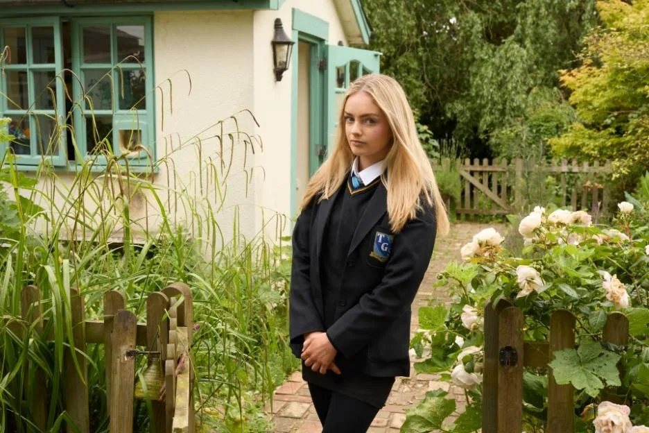 Beatrice Hart, a young blonde woman in a school uniform, stands in a garden.