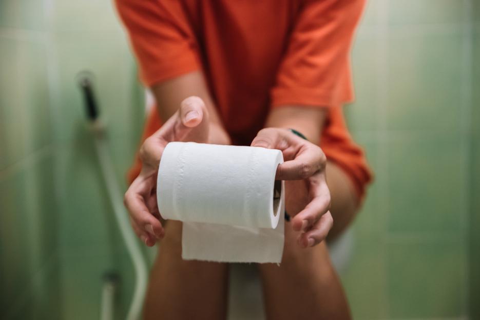 A woman sitting on a toilet holds a roll of toilet paper.