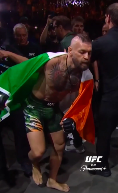 Conor McGregor walking without shoes, with an Irish flag draped over his shoulders, at a UFC event.