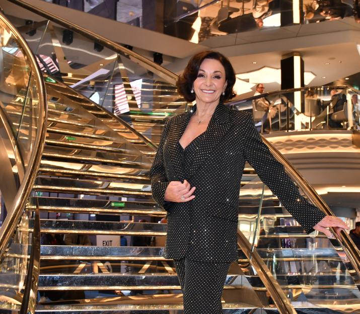 Shirley Ballas in a black suit with sparkling embellishments, standing on a grand staircase.