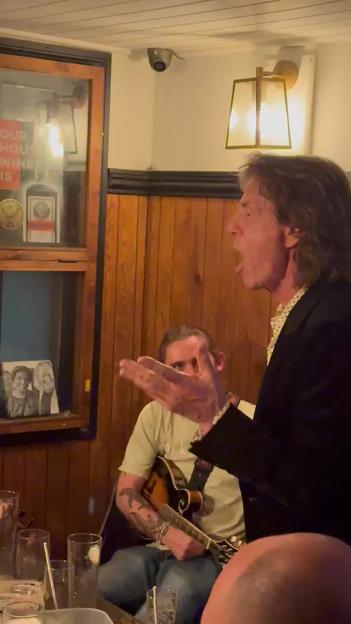Mick Jagger singing in a pub, with a mandolin player behind him.