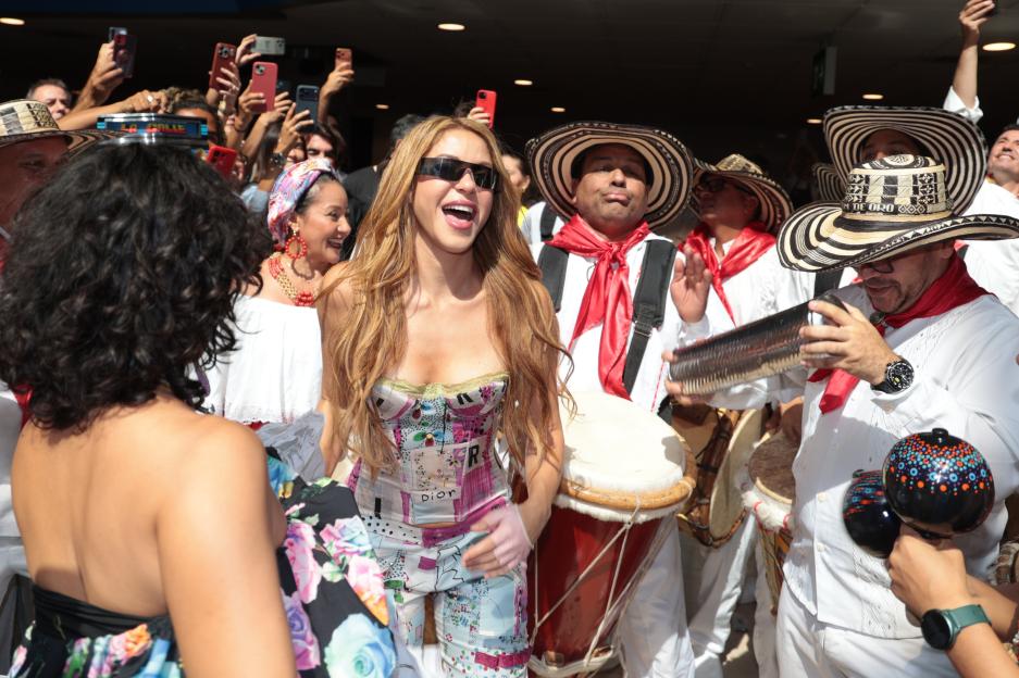 Shakira, wearing sunglasses and a patterned corset and pants, sings and laughs as she is greeted by a band in traditional clothing, playing drums and other instruments, with fans recording on phones in the background.